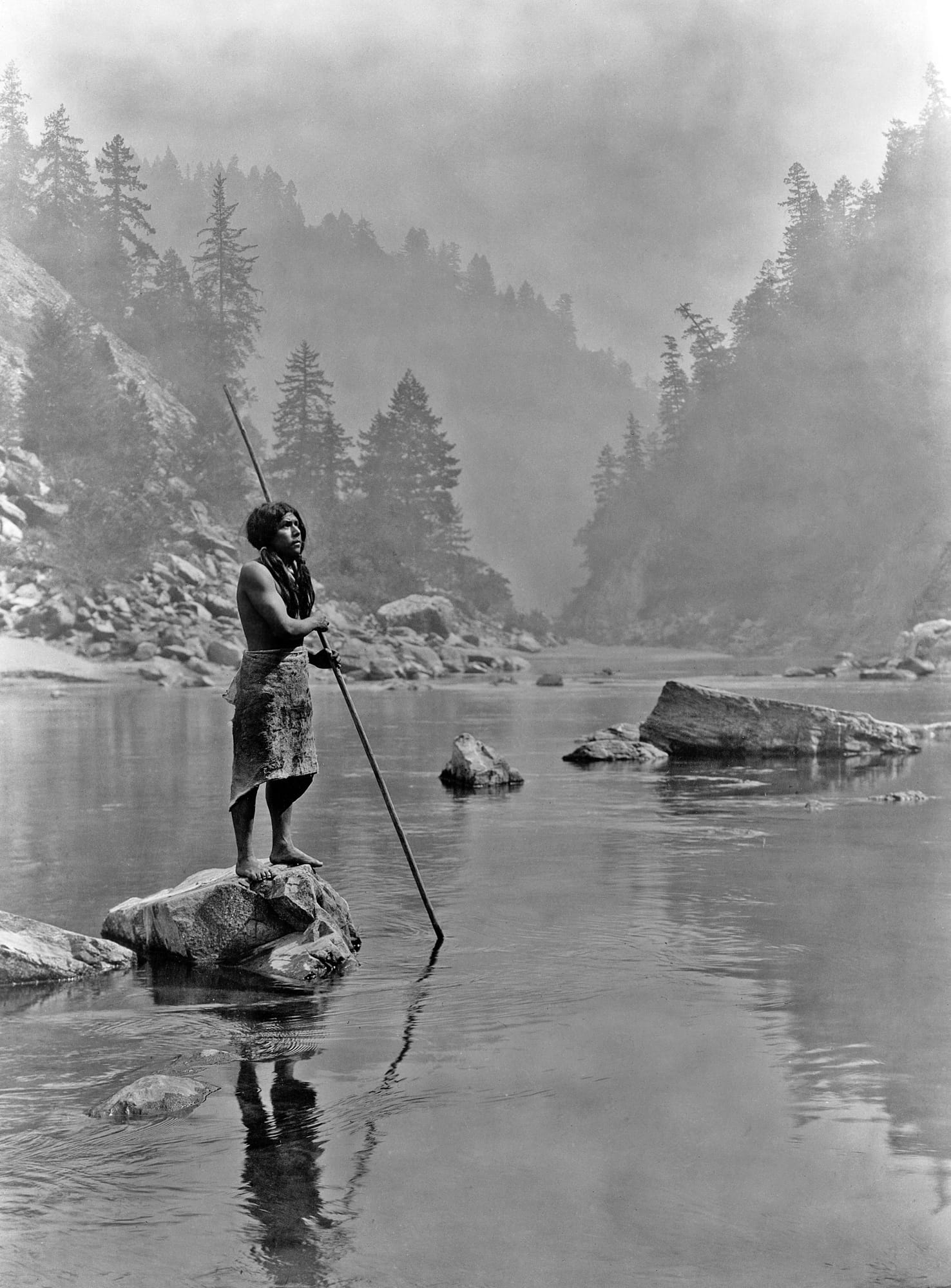 A smoky day at the Sugar Bowl--Hupa. Hupa man with spear, standing on rock midstream, in background, fog partially obscures trees on mountainsides. Published 1924 in The North American Indian / Edward S. Curtis. [Seattle, Wash.]&nbsp;: Edward S. Curtis, 1907-30, Suppl., v. 13, pl. 471.
