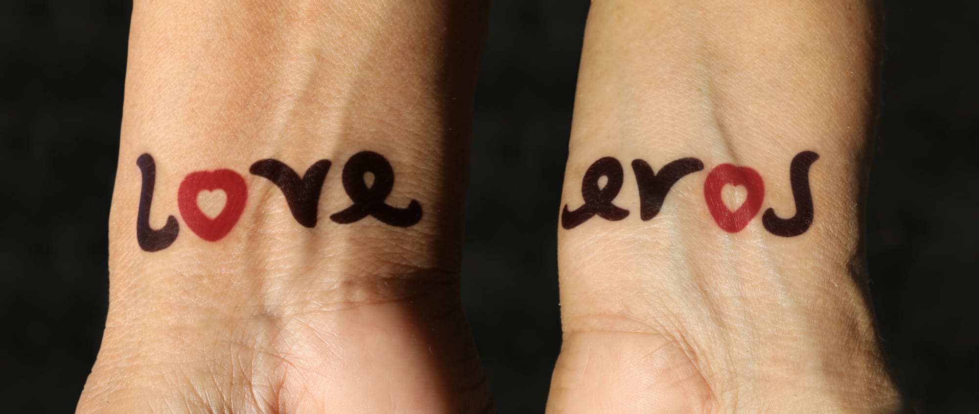 Ambigram tattoo Love / Eros, one word on each wrist. Mirror symmetry (vertical axis). Design with a red heart symbol for the letter O in both words. Eros (concept) is  one of the four ancient Greco-Christian terms which can be rendered into English as "love" (the other three are storge, philia, and agape). Eros refers to "passionate love" or romantic love. The term erotic is derived from eros. Eros has also been used in philosophy and psychology in a much wider sense, almost as an equivalent to "life energy". Decal-style temporary tattoo.