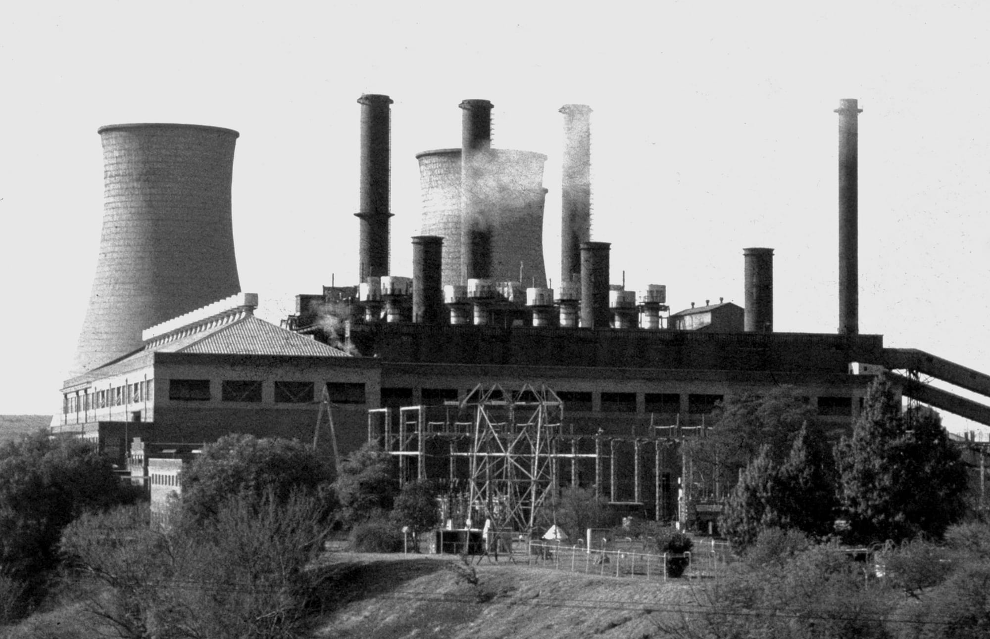 View of Colenso Power Station taken from the railway bridge over the Tugela River in the 1970's. The power station was built in the 1920s and decommissioned in 1985. This photo was digitised in 2014 after the power station had been demolished. 
The river bank is on the left of the picture, the cooling towers in the background, the transformer yard in the foreground and the coal conveyor belts on the right-hand side of the picture.