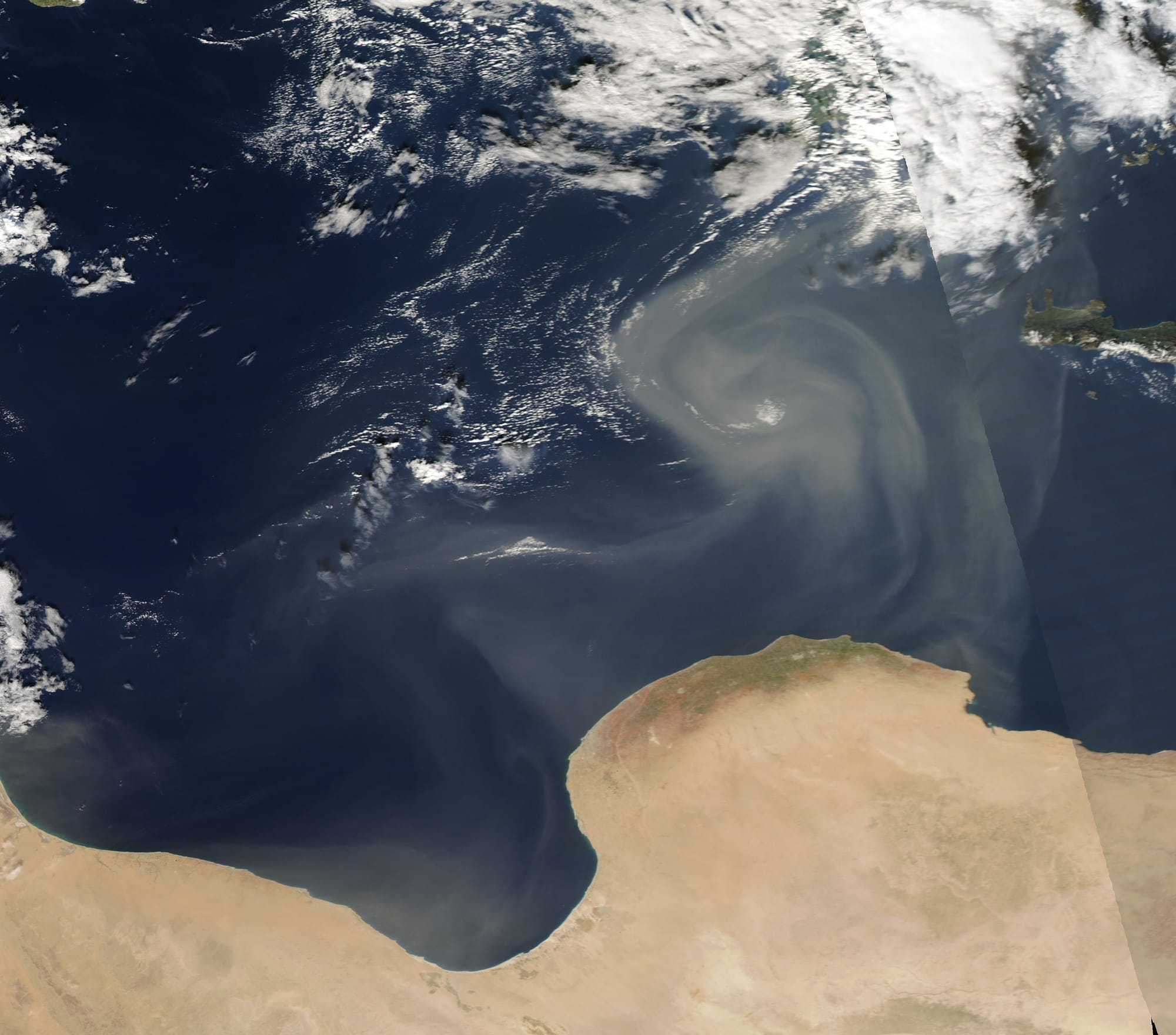 Gusting winds carried Saharan dust over the Mediterranean Sea in mid-November 2022.
On November 17, the Moderate Resolution Imaging Spectroradiometer (MODIS) on board NASA’s Terra satellite acquired a true-color image of the start of a dust storm, with camel-colored sand and dust blown into a swirling pattern between Libya (south) and Greece. Subsequent MODIS images show that clouds of dust have continued to pulse over northern Libya and the Mediterranean through November 19. While the exact source of the dust isn’t clear, it appears to primarily rise from sand seas in northern Libya.

Libya, a country about two-and-a-half times the size of the U.S. state of Texas, is one of the driest countries in the world. According to the United Nations Office for Coordination of Human Affairs, as of 2021, the country is more than ninety-five percent desert, with most of the land barren with flat to undulating plains. Only about one percent of the land is arable, and the country is forced to import about 75 percent of the food needed to meet local needs. OCHA notes that the Great Man-Made River project, which provides 60 per cent of all fresh water used in Libya, draws water from non-renewable aquifers that cannot be recharged by rain. The country is susceptible to floods, sandstorms, dust storm and desertification. The UN notes that Libya is a country living in “fragile circumstances” and it is one of the areas severely vulnerable to the effects of climate change.