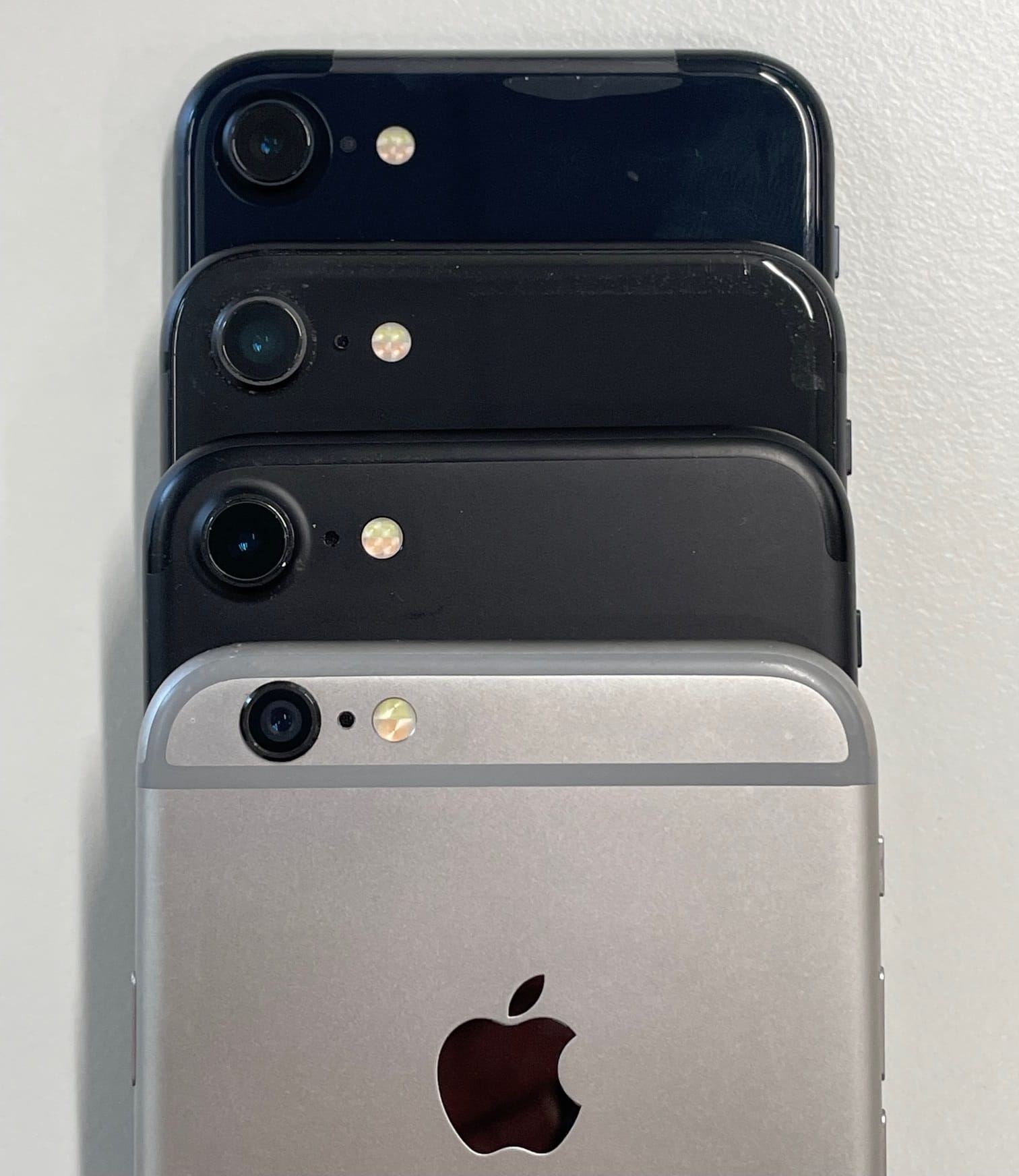 Iphones - models 6, 7, 8 and SE2 - stacked for comparison. These models have nearly identical case shape and dimensions. View of the back side top section, showing: camera, microphone hole, dual-tone LED illuminator. 6 and 7 have aluminium backs whilst 8 and SE have glass.  iPhone 6 is "space grey" colour, others are black or "midnight". The SE 2nd gen is new out of box and still has protective shipping wrapper attached; others are used and showing some wear.