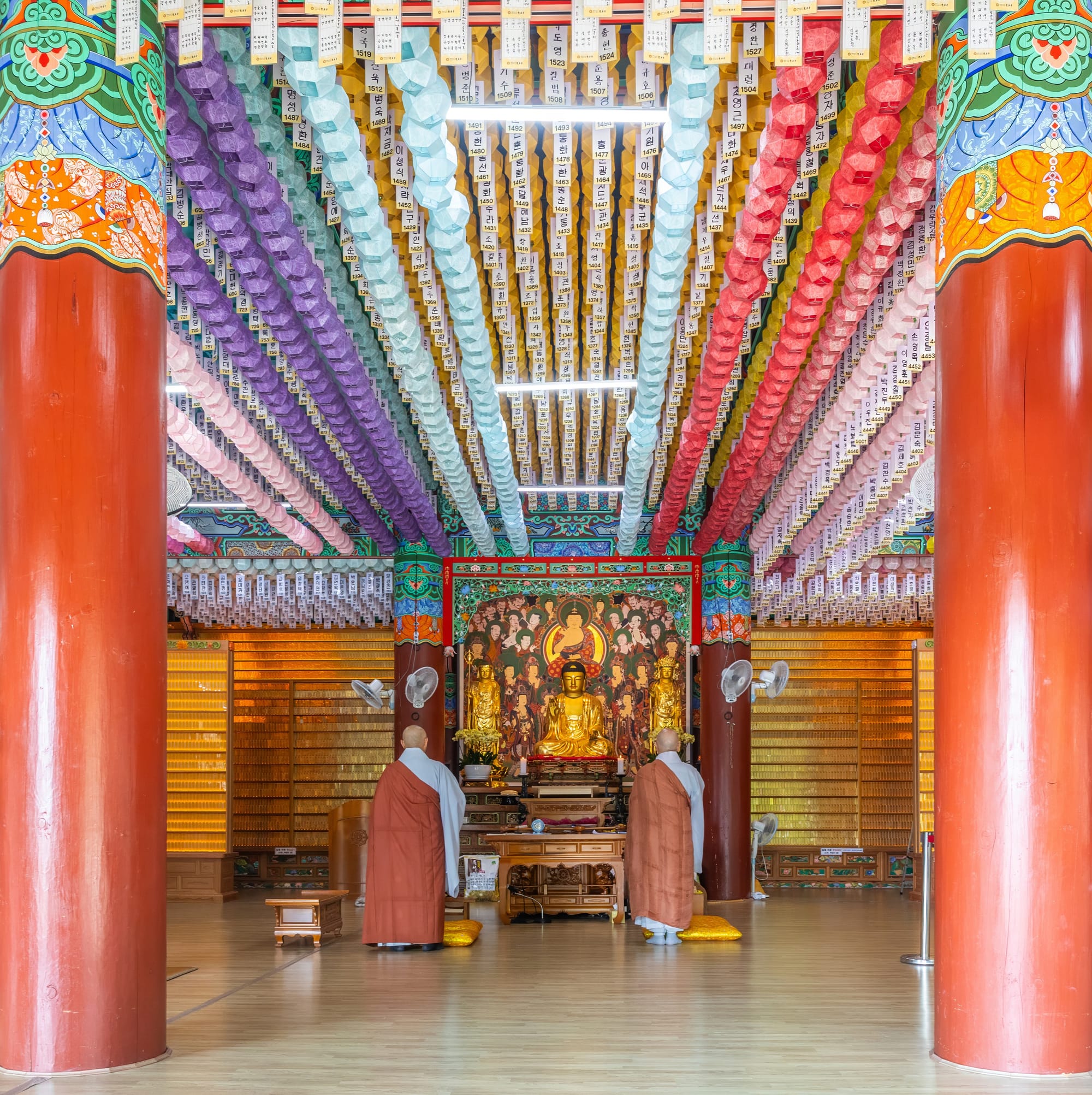 Interior view of Beomeosa Buddhist temple during a religious ceremony, with paper lanterns on the ceiling, red columns, a gold statue of the seated Buddha, and two Buddhist monks seen from behind, in Busan, South Korea.