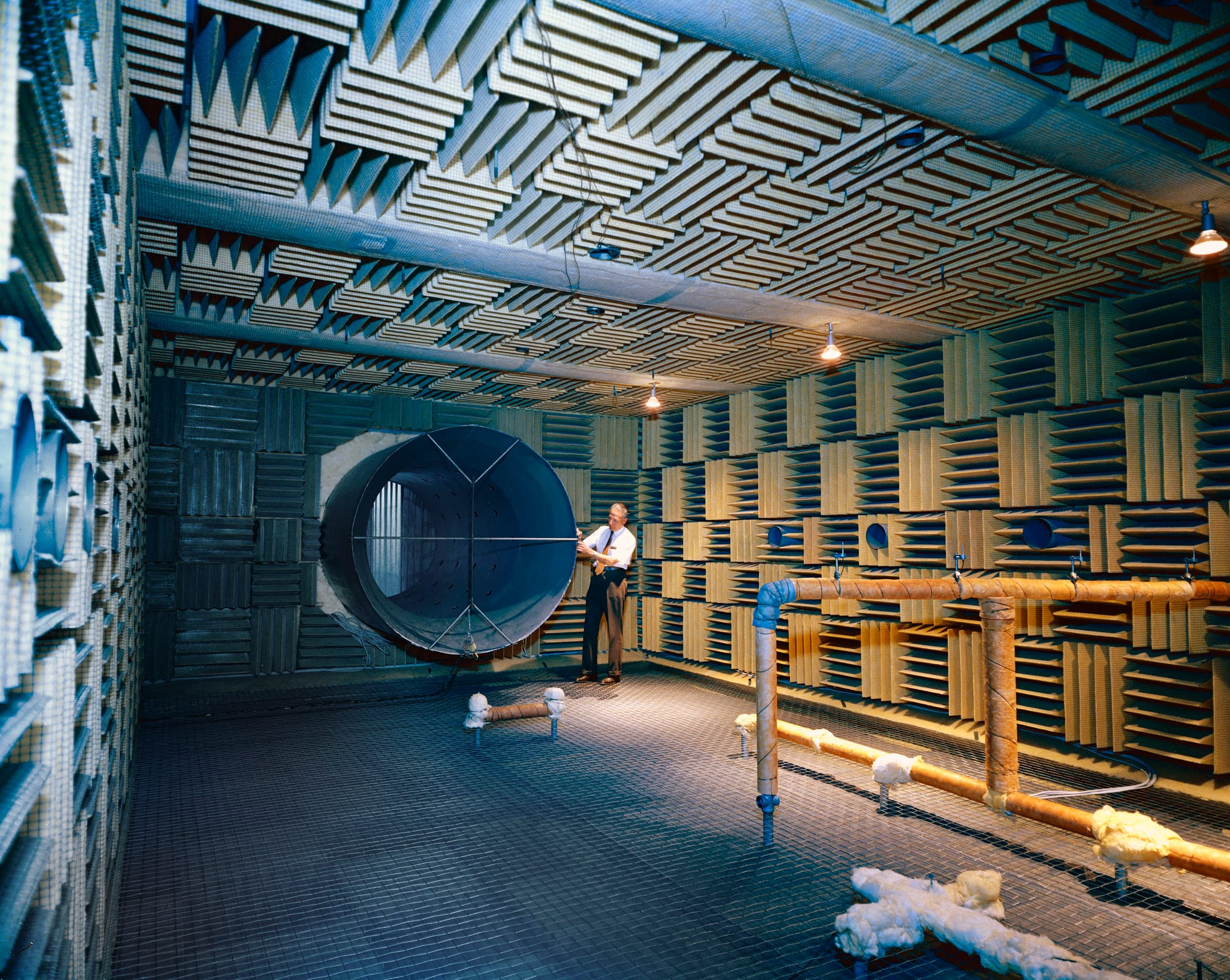 This aerodynamic noise facility, also called an anechoic chamber, was used to study the noise generating mechanisms in supersonic and subsonic jets in the early 1970s. It was in building 57 (which no longer exists) located next door to the wind tunnel that was in building 79 at the time. The large round opening in the wall is an exhaust silencer inlet. Standing next to it is Paul Massier, co-author of a technical report about this chamber. On the right is a support structure for microphones. Fiberglass wedge blocks cover the ceiling and walls, which were mostly reinforced concrete. Spaces were left open to allow for observation windows and instruments to record test data. There were also openings in the walls that allowed air to flow into the chamber to replace the air forced out during tests.
Aug. 30, 1971
Requested by: P. Massier
Photo by: J. Rayle