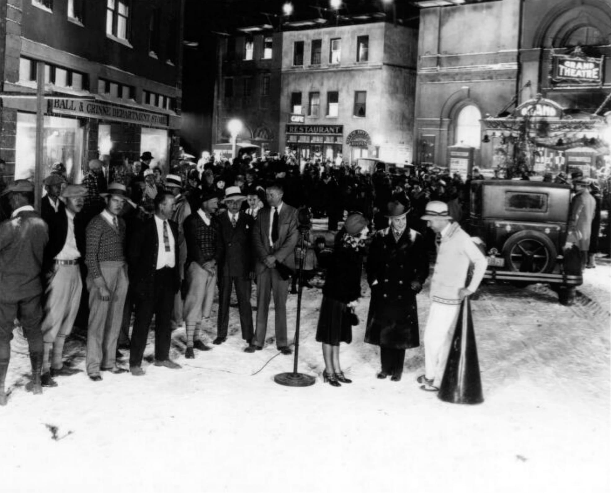 Norma Shearer and Oscar Shaw on the set of Upstage. Text from back of photograph: Back lot at M-G-M, Culver City. Night snow scene - showing the loudspeaker microphone over which I direct the 250 extras, autos & horses. MONTA BELL / NORMA SHEARER / OSCAR SHAW / NICK GRINDE / JOE RAPF