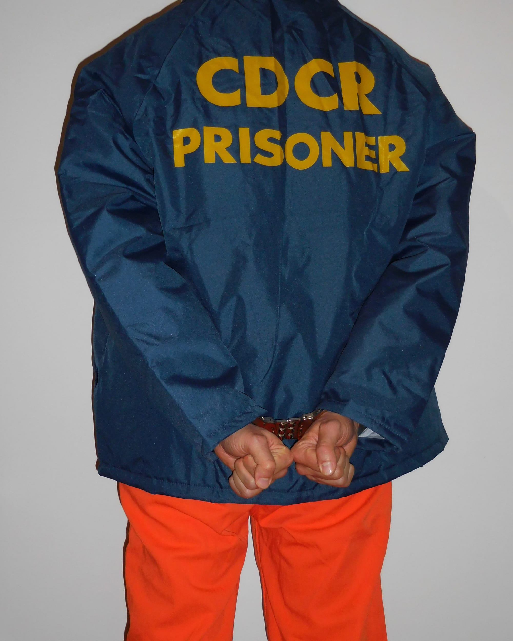 Prisoner of the California Department of Corrections and Reahbilitation wearing orange pants, a dark blue jacket and a pair of hinged handcuffs.