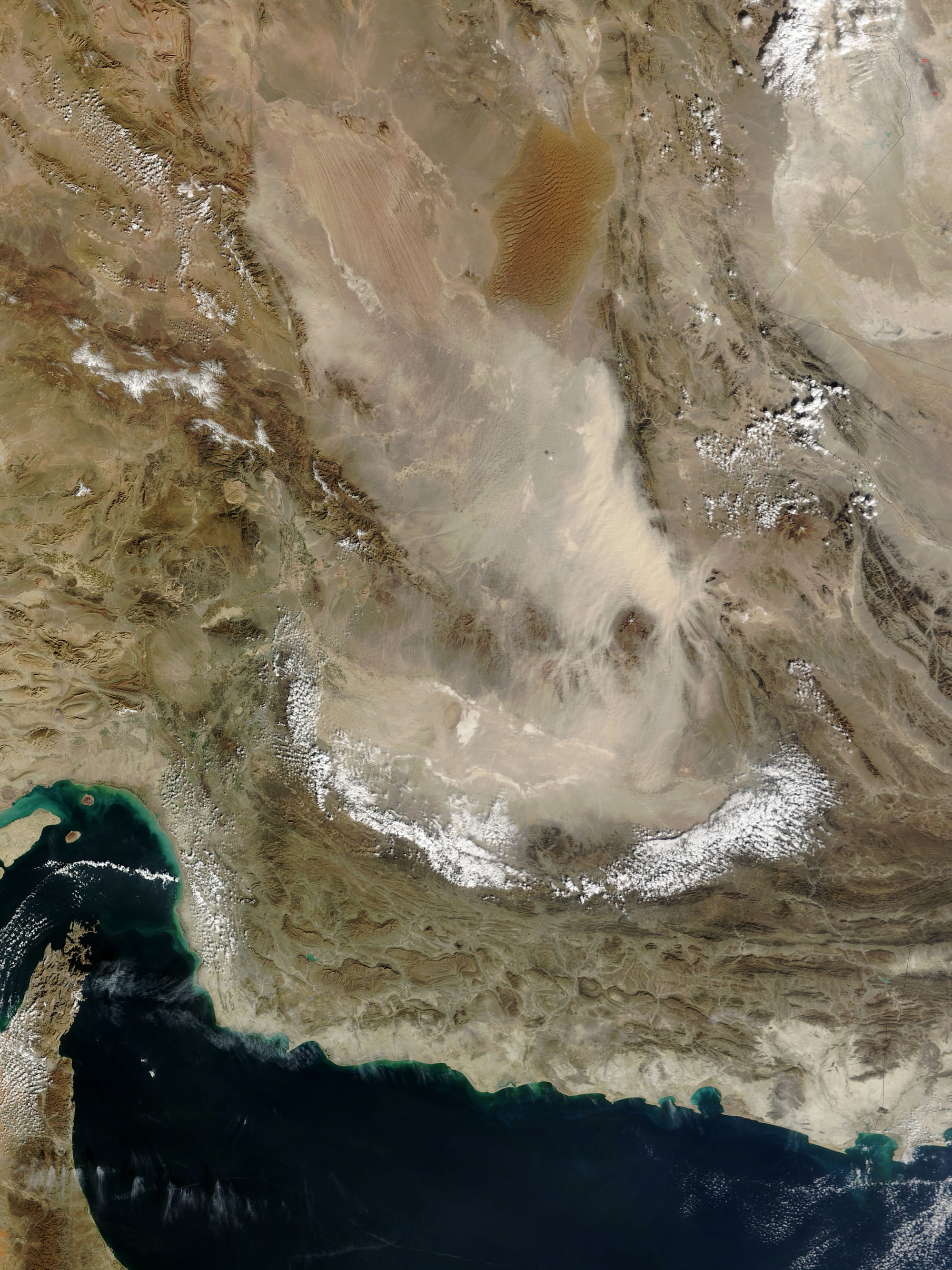 Pale tan dust billows over the Dasht-e Lut in eastern Iran, in this photo-like image from the Moderate Resolution Imaging Spectroradiometer (MODIS) on NASA’s Terra satellite. In the thickest part of the storm, the dust cloud blurs the texture of the land beneath it, revealing instead the texture of the air in waves and lines. The most distinctive waves surround Kuh-e Bazman, a 3,489-meter (11,450-foot) tall volcano, which rises above the dust cloud. The ripples in the dust are a reflection of turbulence created as air flows around the peak. The dust seeps into the valleys of the mountains south and east of the volcano.
The Dasht-e Lut, is a large salt desert in remote eastern Iran. The dust storm blows across the southern desert, leaving clear the distinctive orange dune fields and wind-swept lines in the north.