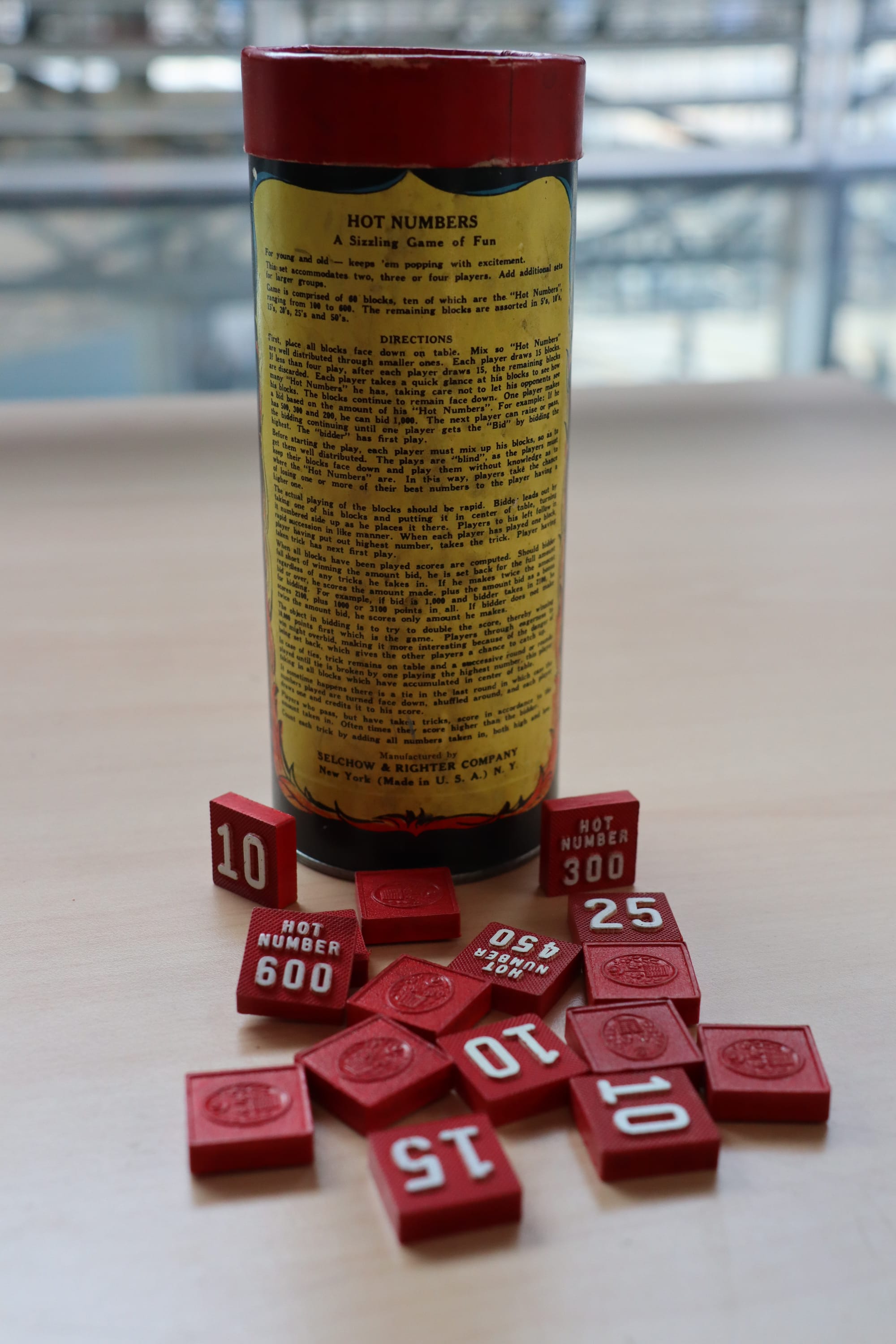 Cette image montre le jeu américain "Hot Numbers". On aperçoit sur la photo le tube de rangement du jeu avec la notice écrite, ainsi que des pièces du jeu en forme de cubes rouge inscrits de différents numéros. Il s'agit d'un jeu d'estimation.