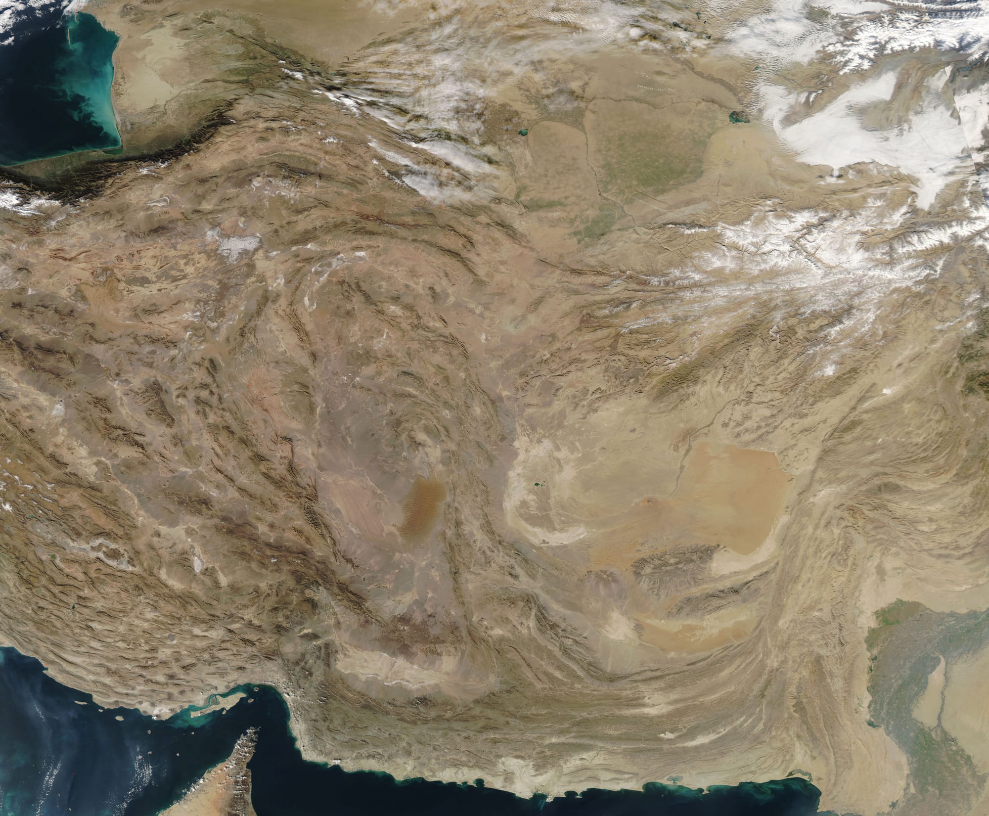 On December 16, 2018, NASA’s Aqua satellite passed over the Middle East, allowing the Moderate Resolution Imaging Spectroradiometer (MODIS) on board to acquire a beautiful true-color image of Iran, Afghanistan and Pakistan.
One notable feature readily identifiable in the west (left) of the image is the upfolding topography of the Zagros Mountains. Beginning north of the Persian Gulf near the narrow Strait of Hormuz, the long mountain range extends about 990 miles (1,600 km), forming the extreme western boundary of the Iranian plateau. The area is actively undergoing crustal shortening as global tectonics move Arabia towards Asia. As a result, the layers of sedimentary rock are slowly folding upward in ridges, somewhat like a carpet folds upward if pushed. In this case, the compressive forces create massive folds – the local relief between the higher mountain ridges and the valleys can be as much as 1200 meters (4,000 feet).

To the east, in southern Afghanistan, an apparently smooth area of orange-tinted tans marks the large Registan Desert. The desert terrain is, indeed, relatively flat, but fails to meet the criteria of “smooth”, at least at ground level. The winds, which blow consistently form the same direction, causes sand to pile up in crescent-moon shaped dunes, whose thin tips point in the direction of the wind flow. Called barchan dunes, these dunes will migrate across the desert surface as sand grains on the crest of the dune are toppled by the wind and spill down the leeward face. As the pile of sand on the leeward side of the dune gets larger, gravity eventually topples the pile, moving the dune’s leading edge slowly forward. The movement is slow but steady – some barchan dunes can migrate between 8 and 15 feet each year.