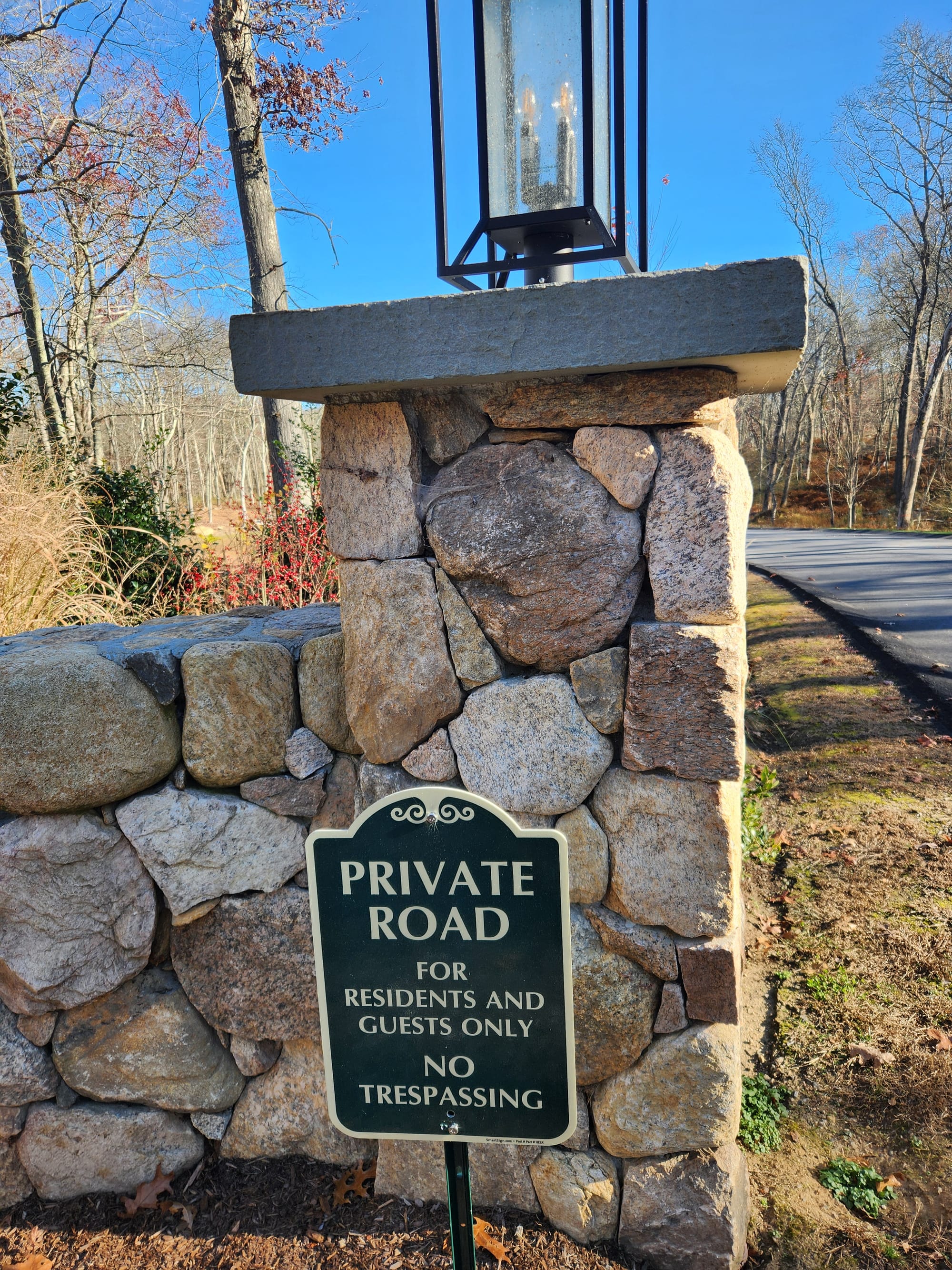 A decorative stone pillar with a lantern at the entrance to a private road. A sign indicates restricted access with the text: "Private Road for Residents and Guests Only - No Trespassing." Located in a wooded area on a clear autumn day.