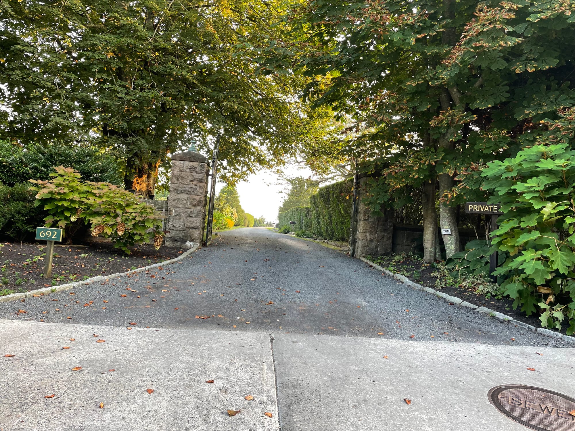 The photo depicts a residential driveway entrance during the daytime. The entrance is marked by a stone pillar on the left with the house number "692" displayed on a small green sign. On the right, another stone structure is partially obscured by lush greenery and a sign that reads "PRIVATE," indicating that this is a private driveway. The asphalt driveway curves gently to the right, leading towards an area not visible in the photo. Overhanging branches from mature trees create a canopy over the entrance, and the green foliage is dense on both sides. There are fallen leaves scattered on the driveway, suggesting an early autumn season. The sky appears clear with soft lighting, likely indicating either morning or late afternoon. There is a manhole cover in the foreground on the sidewalk, and the general atmosphere is peaceful and secluded.