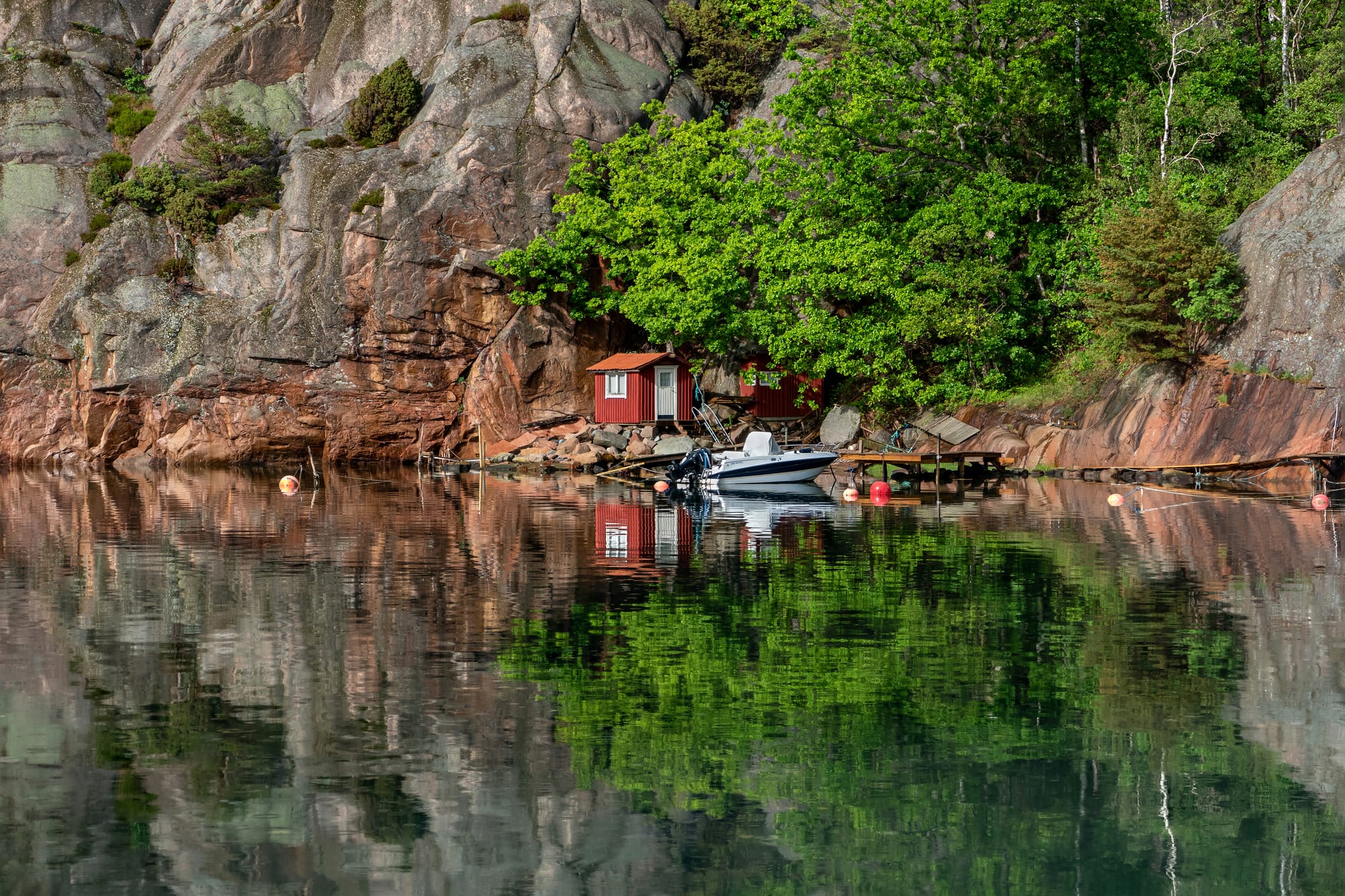 Two Falu red fishing huts and a motorboat by the cliff in Rågårdsdal, Lysekil Municipality, Sweden. A big oak (Quercus robur) fills up most of the lower part of the crevasse. Photo taken while it was still raining, making patterns on the water surface and enhancing the color of the cliffs.