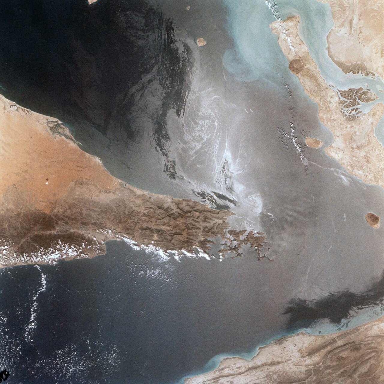 Portions of Oman, The United Arab Emirates and Iran are seen at the Strait of Hormuz (26.0N, 56.0E) in this view. A number of ship wakes can be seen in the area of the strait. Sunglint in the Persian Gulf to the northwest, accentuates the complex currents and oil slicks, seen as dark toned blue streaks on the surface. Qeshm Island, just off the coast of Iran, can be seen on the edge of the photo.