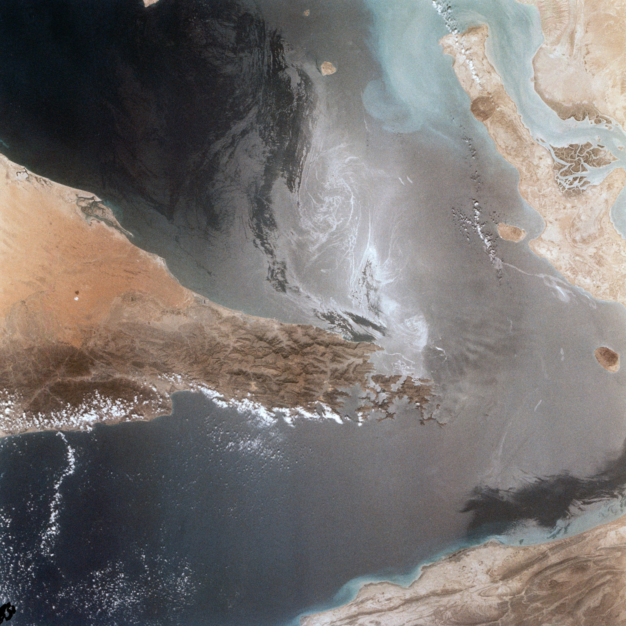 Portions of Oman, The United Arab Emirates and Iran are seen at the Strait of Hormuz (26.0N, 56.0E) in this view. A number of ship wakes can be seen in the area of the strait. Sunglint in the Persian Gulf to the northwest, accentuates the complex currents and oil slicks, seen as dark toned blue streaks on the surface. Qeshm Island, just off the coast of Iran, can be seen on the edge of the photo.