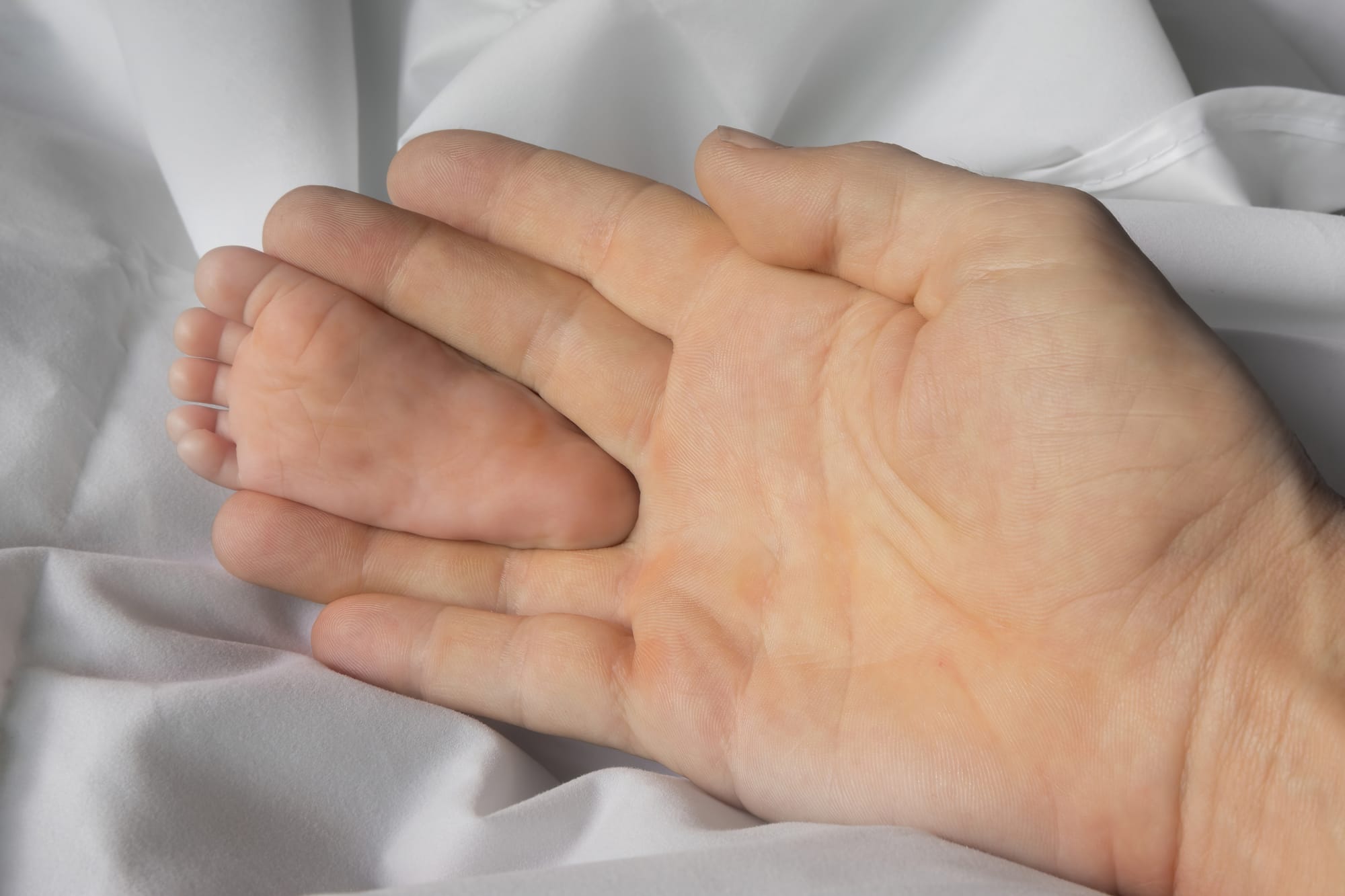 Sole of foot of a one month old infant, held between the middle finger and the ring finger of an adult hand, for size comparison, on a white bed sheet, in Don Det, Si Phan Don, Laos.